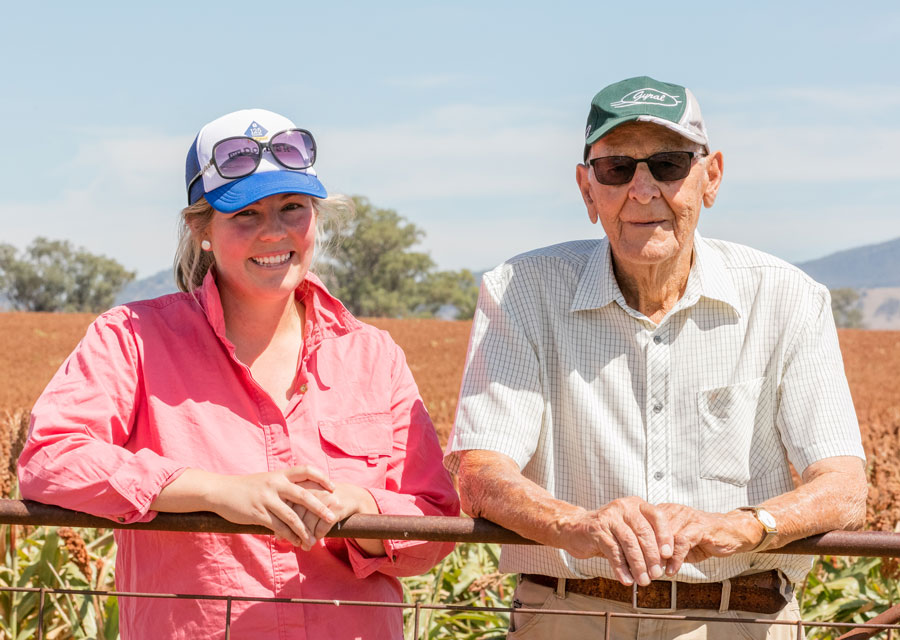 A photo of Emily and Jim leaning on a farm gate and smiling at the camera with a sorghum crop behind them.