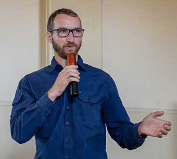 Mat Dunn has glasses on and a beard and he is wearing a dark blue shirt. He is speaking into a microphone