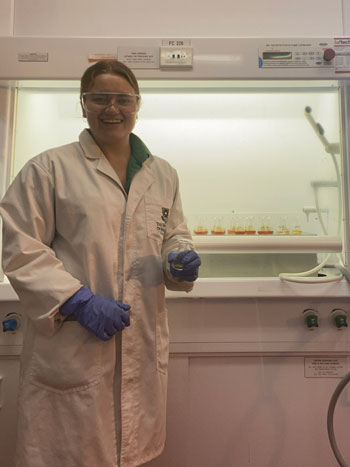 A photo of Megan Hunter in a laboratory wearing a lab coat, safety glasses and blue gloves holding a flask and smiling at the camera.
