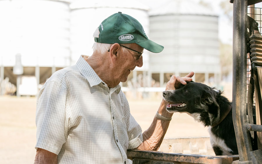 A photo of Jim Hombsch patting the head of a dog who is in the back of a ute. He is facing sideways from the camera and gently smiling at the dog.