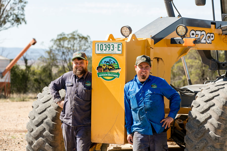 Matt and Brad Jackson posing either side of a John Deere grader with a Jackson Brothers logo on the front of it. They are both smiling at the camera. 