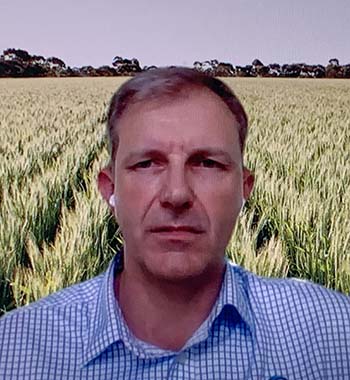 A man in a blue checked shirt with earbuds in against a backdrop of green flowering wheat.