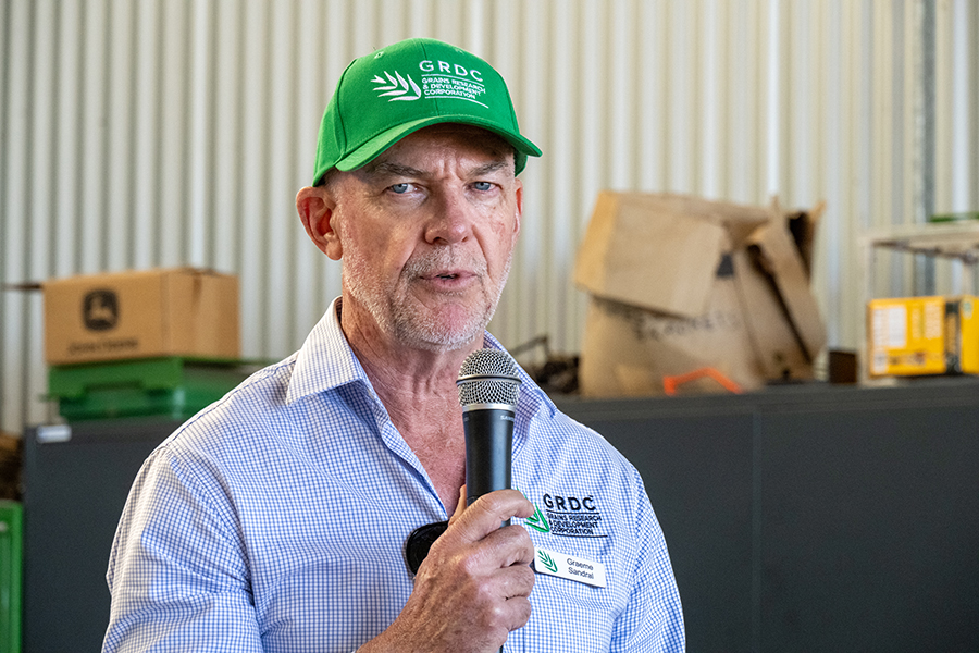 Graeme Sandra is wearing a green GRDC cap and blue checked GRDC shirt. He is holding a microphone and looking at a the camera as he speaks to the audience in Charlie Baldry's shed.