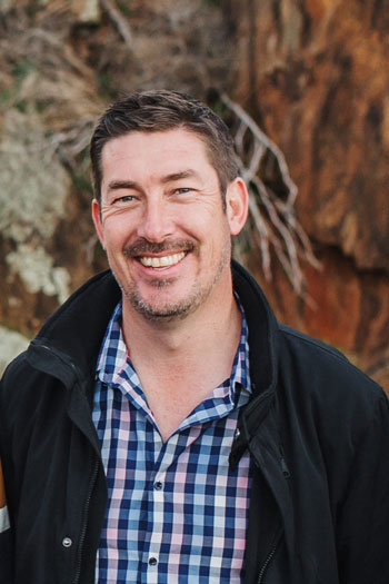 A portrait photo of Paul McGorman smiling at the camera in front of a blurred reddish rock background.