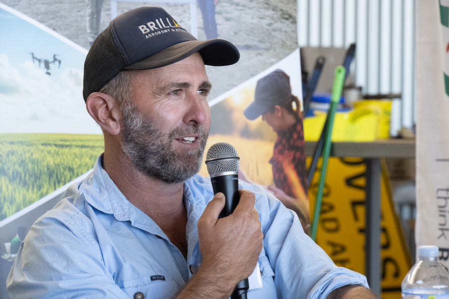 Rohan Brill is wearing a salt and pepper coloured beard, dark blue Brill Ag cap, and blue Brill Ag shirt. He is holding a microphone and looking at the field day audience.