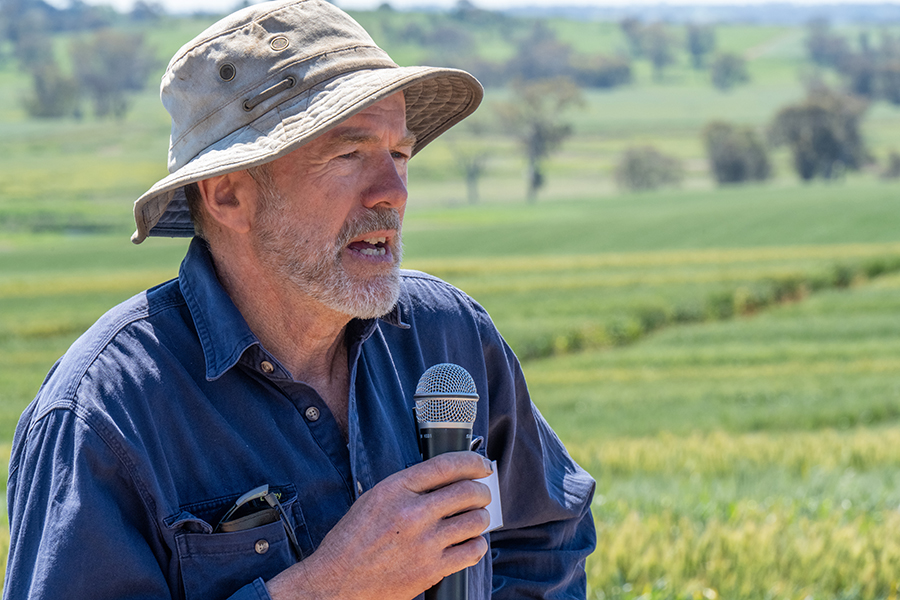 Charlie Baldry is holding a microphone as he speaks to the field day audience in one of his paddocks where the GRDC Hyper Yielding Crops trials are located. He has a brown bucket hat a short beard and dark blue workshirt on.