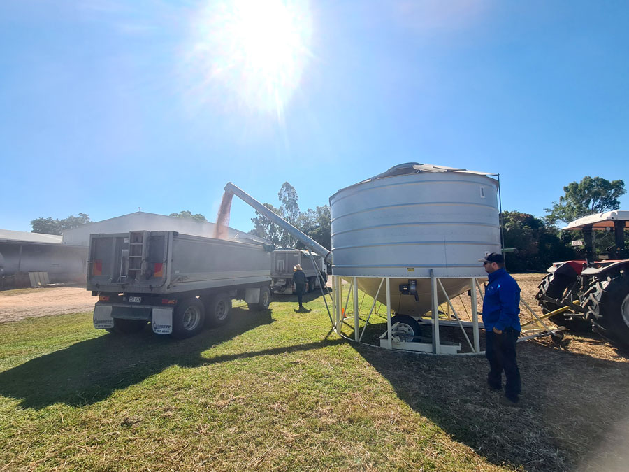 A photo of a small silo being emptied into a truck while a man in a blue long sleeved collared shirt and black cap watches from next to the silo.
