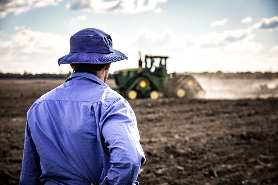 A photo of Ben Taylor in a paddock with his back to the camera as he watches a grader just out of focus in the middle distance. 