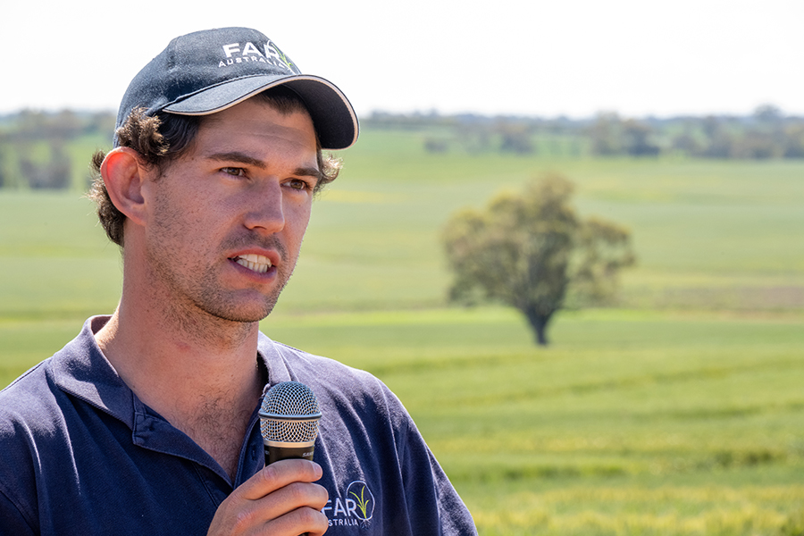 Tom Price is wearing a dark blue FAR Australia cap and t-shirt. He is holding a microphone in the wheat trial paddock and looking at the audience. He has brown short hair and a very short beard.
