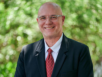 Photo of man with glasses and in a suit standing in front of out of focus trees