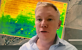 A man with blonde hair and a blue shirt talking in a webinar in front of a background that looks like a coloured yield map.