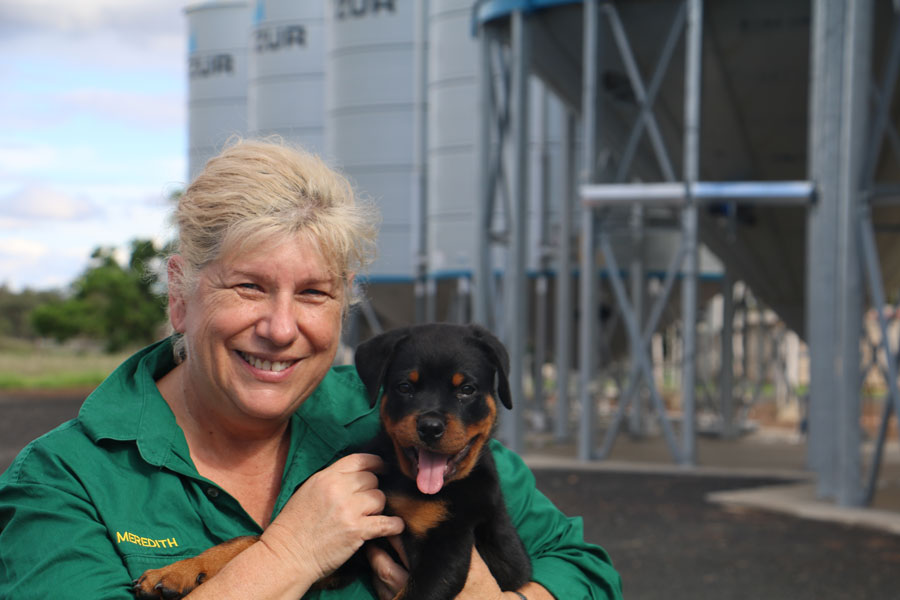 A bust up photo of Meredith holding a black and tan kelpie puppy in front of a row of large silos. They are both smiling at the camera.