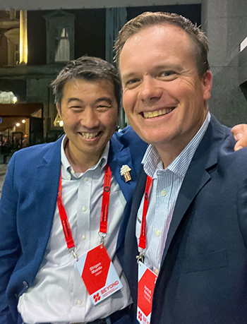 Shigeo and Jarrod at a conference wearing dark blue coats and light blue shirts with red name tags.