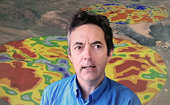 A man in a blue shirt with brown hair speaking to a webinar in front of a precision farming map with red, blue, green and blue colours.