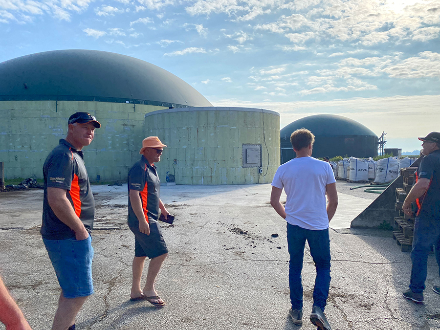 three Wallup Ag Group members in their grey and orange t-shirts with shorts with Maurits te Braake in white t-shirt and blue jeans. In the background are digesters: two domed green structures where organic waste from the dairy is pumped for methane production.