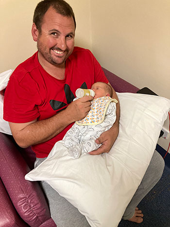 Photo of Robert Pocock holding his newborn baby daughter and feeding her milk from a bottle.