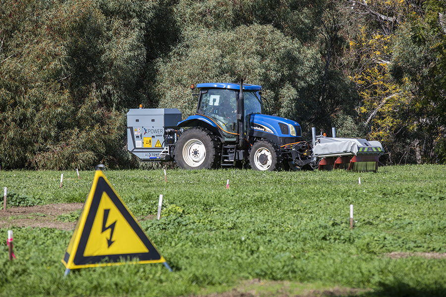 A blue tractor fitted with the AGXTEND XPower electric weed control. There is a box on the back hitch halfway up to the tractor's roof and an attachment on the front of the tractor that looks like it has mudflaps on it.