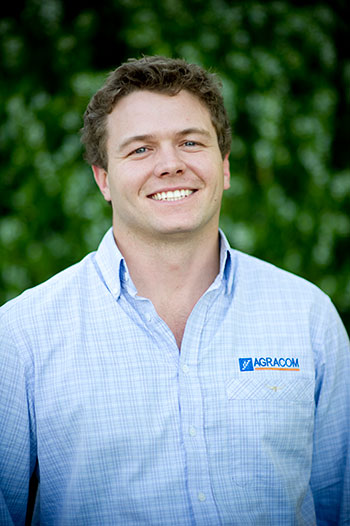 Gordon McNeil is smiling and in a blue checked Agracom shirt. He is standing in front of a green blurred bush.